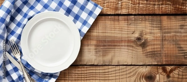 Fototapeta White plate on wooden table with blue checkered napkin. Flat lay, top view, with ample copy space image.