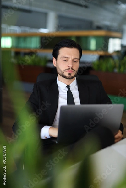 Obraz A man in a suit is sitting at a desk with a laptop open. He is wearing a tie and he is focused on his work. Concept of professionalism and concentration