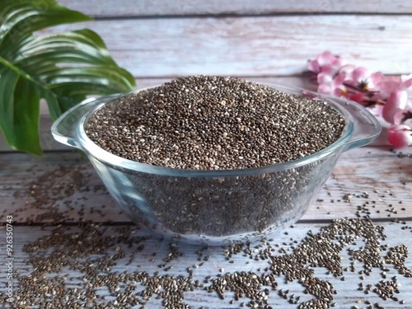 Obraz Clear Bowl of Black and White Chia Seeds on Wood, with Glossy Texture, Scattered Seeds, and Pink Flowers Adding a Decorative Touch in the Background.