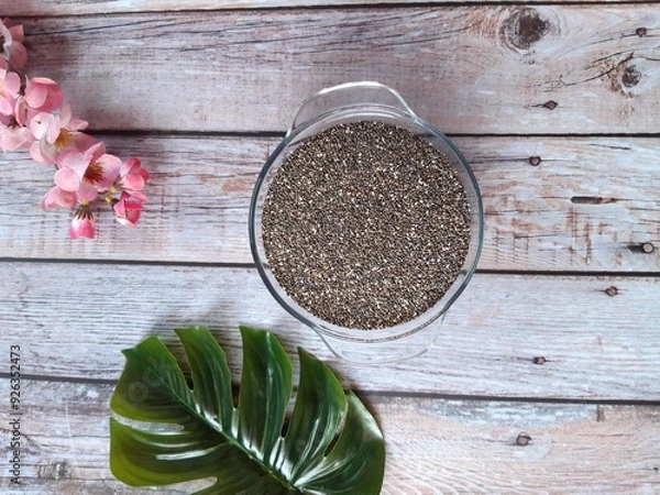 Obraz Glossy Chia Seeds in a Glass Bowl on Wooden Surface, Accompanied by Scattered Seeds and a Decorative Touch of Pink Flowers in the Background.