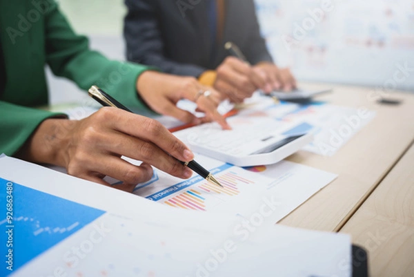 Fototapeta Asian business team engages in a collaborative meeting, financial charts at a desk. Dressed in formal suits, they brainstorm creative ideas to enhance company performance and team cohesion.