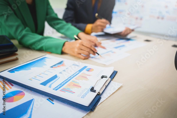 Fototapeta Asian business team engages in a collaborative meeting, financial charts at a desk. Dressed in formal suits, they brainstorm creative ideas to enhance company performance and team cohesion.