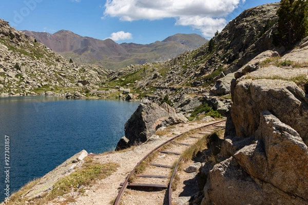 Fototapeta Abandoned wagon tracks on the side of a blue water pond with mountains in the background
