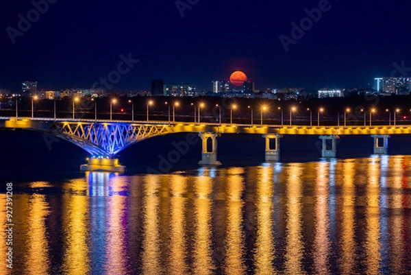 Fototapeta The full moon rises at night. The bridge over the Volga River between the cities of Saratov and Engels, Russia.