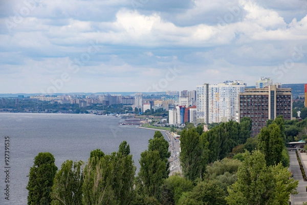 Fototapeta Volga River embankment in Saratov, Russia. Clouds in the sky. Trees and various buildings.