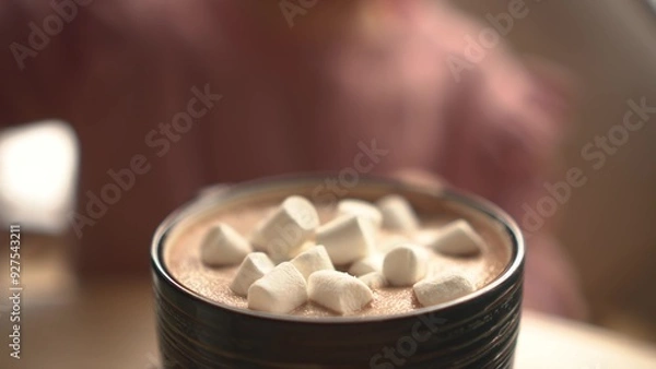 Fototapeta Girl sprinkling marshmallows into a cup of hot chocolate on a green saucer, placed on a wooden table, with a cozy cafe background. Close-up.