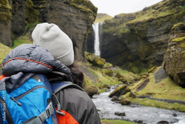 Fototapeta Hiker man arriving at a landscape with impressive waterfall in Iceland. back view. dressed in the mountain suit and hat