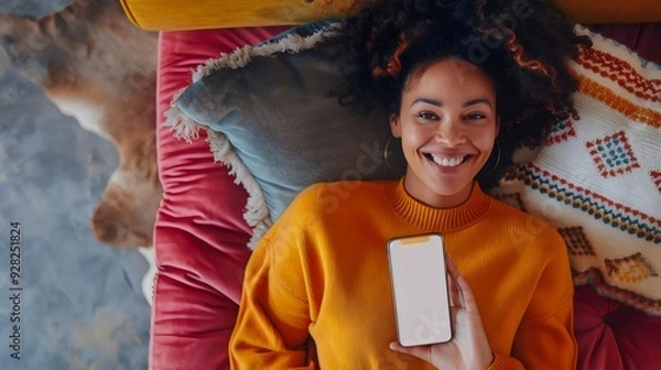Fototapeta A cozy scene of woman lying on a comfortable couch wearing a bright orange sweater while holding a smartphone, surrounded by colorful cushions and a relaxed atmosphere, perfect for unwinding.