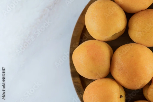 Fototapeta Ripe apricots on a wooden plate, top view. Fresh natural apricots, delicious summer dessert, on marble countertop, close-up, high resolution.
