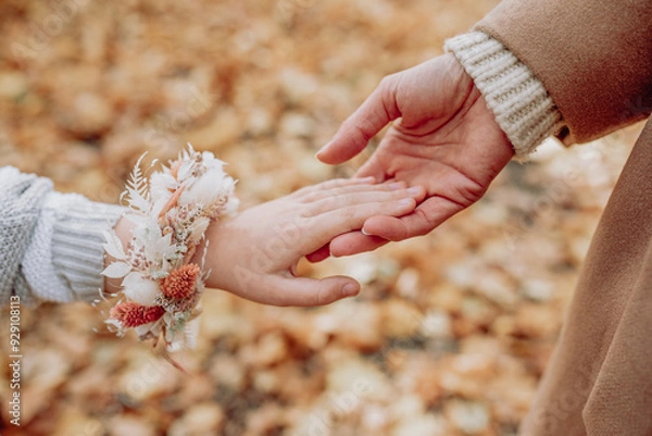 Fototapeta Close-up of two hands touching, one child wearing a floral bracelet and the other an adult, set against a backdrop of autumn leaves, capturing a tender, intergenerational moment filled