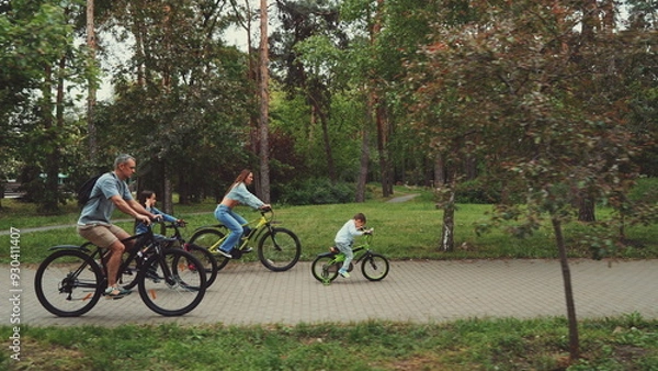Fototapeta Family enjoying bike ride in park, with parents and children cycling together on paved path surrounded by trees. Concept of family bonding, outdoor activities, and healthy lifestyle