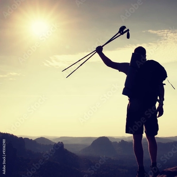 Fototapeta Silhouette of tourist with poles in hand. Sunny daybreak in rocky mountains. Hiker with sporty backpack, view  point above misty valley.