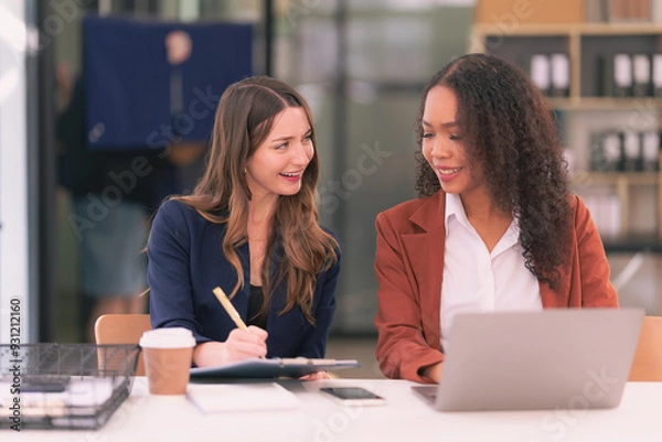 Obraz Two young businesswomen work together on laptops while talking about panning, reading documents and writing notes while discussing a new project.