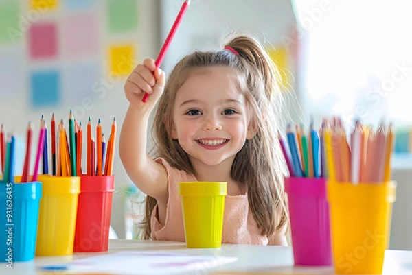 Obraz Photograph of a happy girl holding colored pencils in her hands, sitting at a table in a school classroom setting.