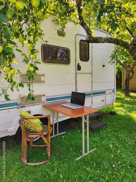 Fototapeta Serene outdoor office setup with a laptop on a wooden table beside an RV and under apple trees.