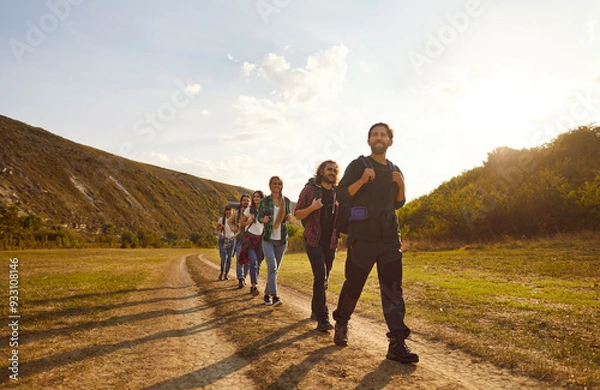 Fototapeta Young happy people tourists with large backpacks hiking in nature walking in a row in the mountains. Smiling friends having trekking tour outdoors. Adventure, travel and tourism concept.
