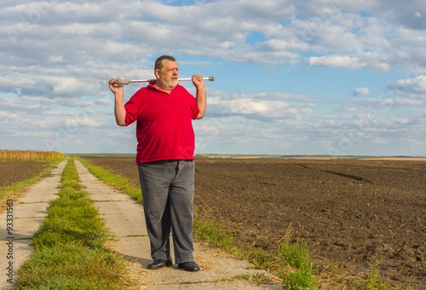 Fototapeta Senior man with walking stick standing on a country road and looking into the distance