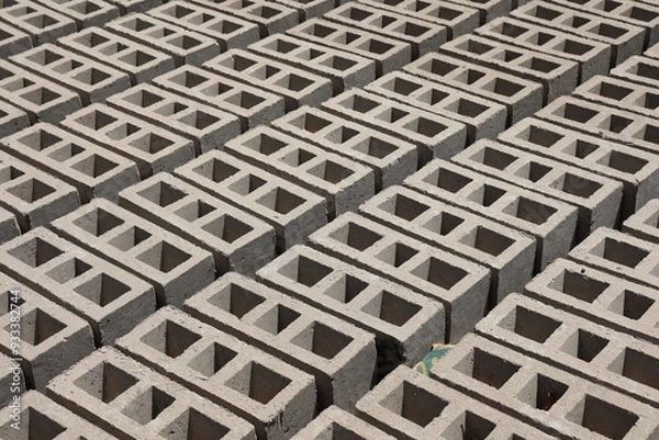 Obraz Rangées de parpaings creux gris en ciment, formés de trois alvéoles, et séchant sur le sol. Matériaux de construction constitués de blocs alignés formant une surface rugueuse. Photo prise au Sénégal.