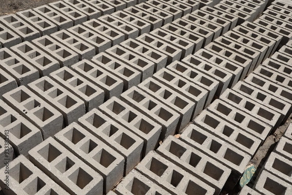Obraz Rangées de parpaings creux gris en ciment, formés de trois alvéoles, et séchant sur le sol. Matériaux de construction constitués de blocs alignés formant une surface rugueuse. Photo prise au Sénégal.
