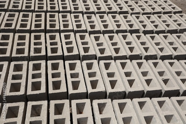Obraz Rangées de parpaings creux gris en ciment, formés de trois alvéoles, et séchant sur le sol. Matériaux de construction constitués de blocs alignés formant une surface rugueuse. Photo prise au Sénégal.