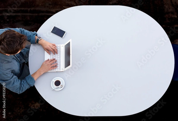 Fototapeta Grey Office Round Table and Man Working on Computer Top View Casual Clothing Typing on Keyboard with Marketing Chart on Screen Smart Phone and Cup of Coffee aside