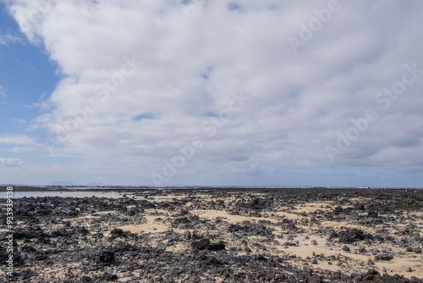 Fototapeta White sand beach with black rocks of volcanic origin. Mountains in the background and sky with big white clouds. Caletón Blanco, Lanzarote, Canary Islands, Spain