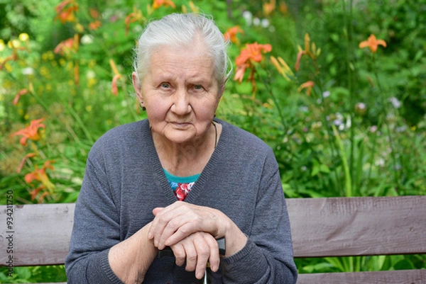 Fototapeta Portrait of a sad elderly woman, 80 years old, with a cane in her hands, sitting on a bench in the park.