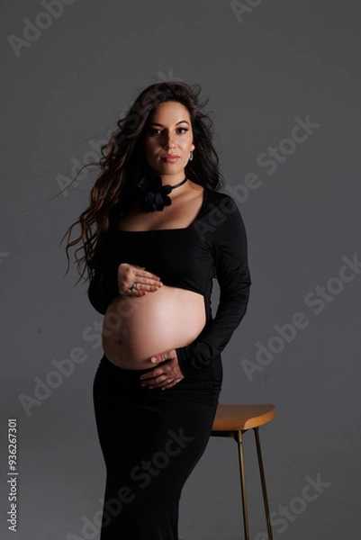 Fototapeta A beautiful pregnant woman with long dark hair with styling and makeup. A brunette waiting for a baby, sit on high chair in a long black dress. Grey background in the studio, fan.