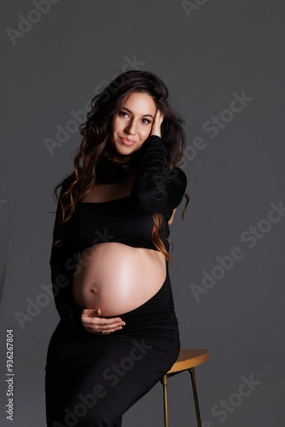 Fototapeta A beautiful pregnant woman with long dark hair with styling and makeup. A brunette waiting for a baby, sit on high chair in a long black dress. Grey background in the studio, fan.