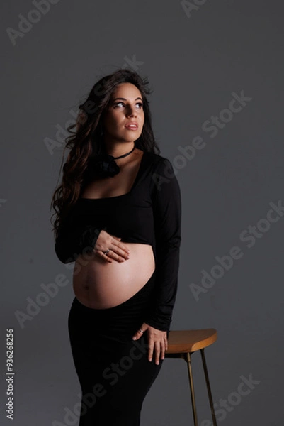 Fototapeta A beautiful pregnant woman with long dark hair with styling and makeup. A brunette waiting for a baby, sit on high chair in a long black dress. Grey background in the studio, fan.