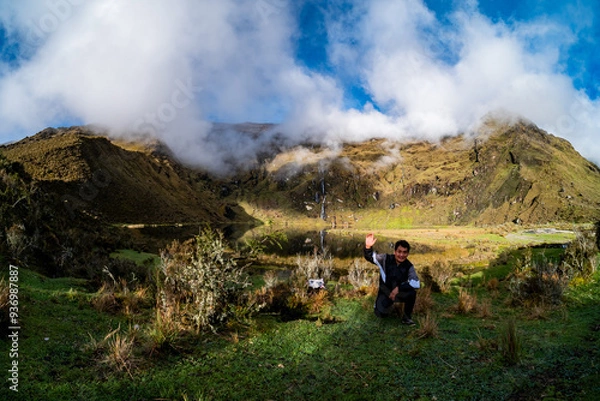 Obraz Hispanic traveler waving while crouching in a mountainous valley, surrounded by green hills and low clouds, with a waterfall and a partially clear sky in the background.