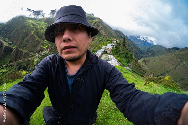 Obraz Selfie of a Hispanic traveler in the Andean mountains, wearing a dark hat and black jacket, with a small rural village and green hills under a cloudy sky in the background.