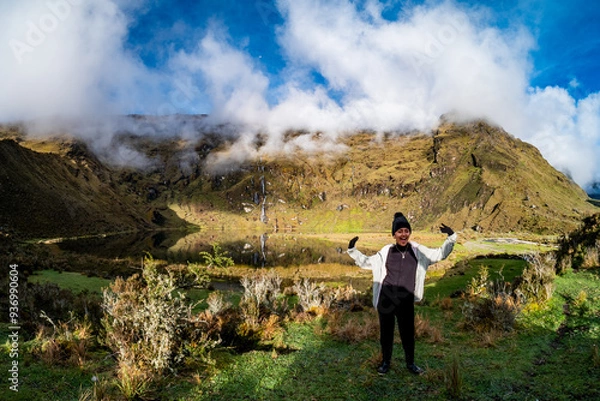 Obraz Young Latina wearing a black cap and beige jacket, waving while crouching in a green mountainous valley, with low clouds, a waterfall, and a partially clear sky in the background.