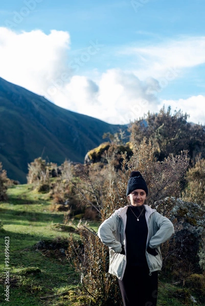 Obraz Portrait of uncomfortable latin woman with arms on waist wearing black beanie and beige jacket against green hills background