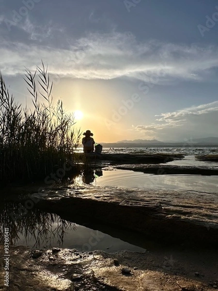 Fototapeta Sunset on Lake Garda on a rocky beach with golden reflections on the water and the sihlouette of a girl at dusk looking at the horizon