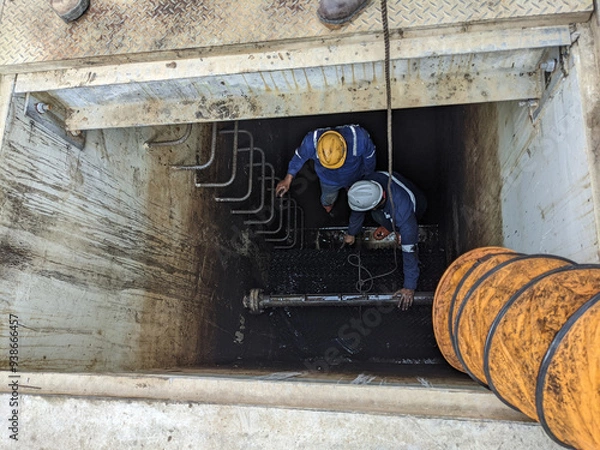 Obraz Workers Inspecting a Tank. Two workers wearing safety gear and hardhats are repairing pipes in a deep trench, showing a common scene of industrial work and maintenance.