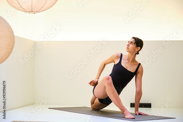 Fototapeta A woman is doing yoga in a studio with a white wall. The room is dimly lit and has a peaceful atmosphere.