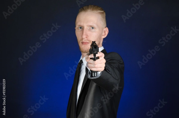 Fototapeta Firearms and security topic: a man in a black suit holding a gun on a dark blue background in studio isolated