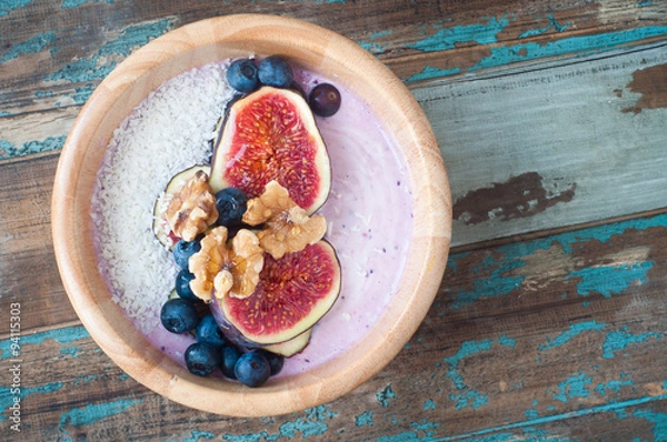 Obraz Smoothie bowl of blueberry and strawberry blended with kefir yogurt and topped with fresh fig, blueberries, walnuts and coconut. Served in a wooden bowl on a rustic wooden table.