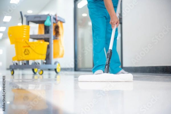 Fototapeta A mop operator in a blue uniform cleans new epoxy floor protection suits in an empty hospital warehouse.