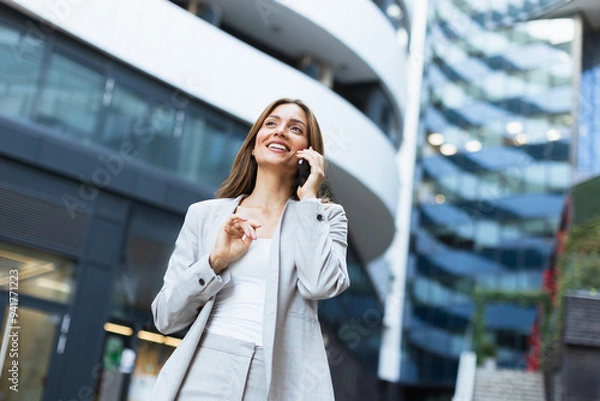 Fototapeta Woman in a light gray suit happily talking on her phone while walking past contemporary architecture in a bustling urban area