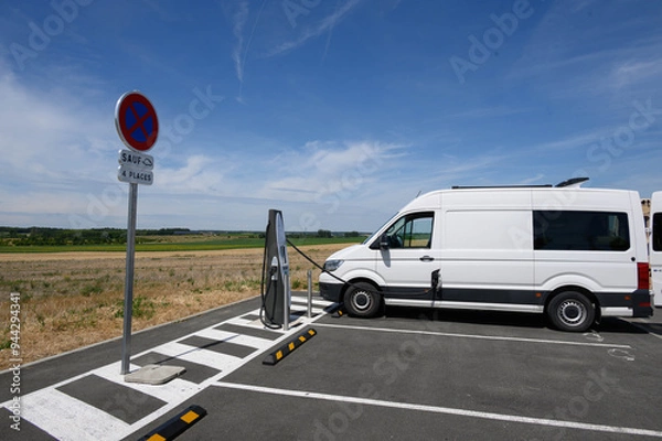 Obraz Side view of an electric van charging at a charge station in the middle of the fields. A sign indicates no parking.  A pictogram authorizes electric vehicles.