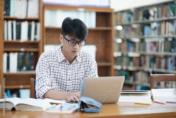 Fototapeta Young Asian male sitting inside a library alone doing research. Man working on a project. Young man doing research for a case.
