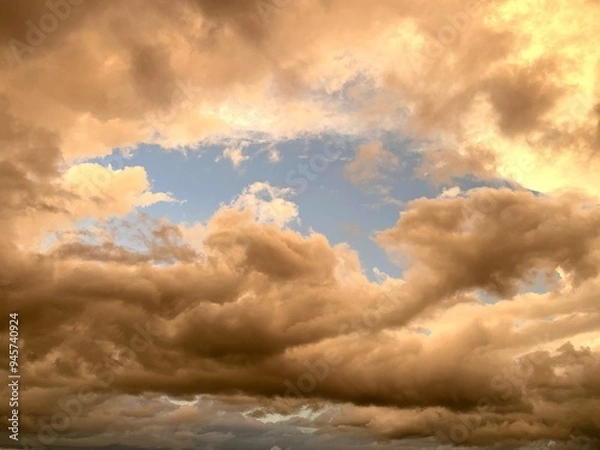 Obraz Fluffy clouds with a yellow tone due to the lighting and a small patch of blue sky shinning through.
