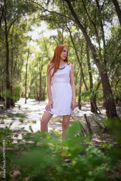 Fototapeta Redhead girl in the white dress posing in the forest thicket.
