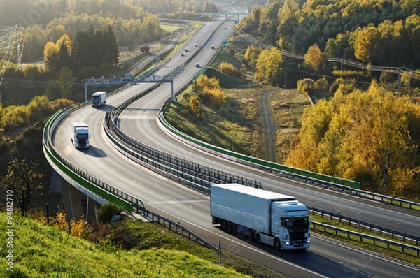 Fototapeta Asphalt highway with electronic toll gates in autumn woodland. Three trucks on the road. The bridge spanning the valley. View from above. Sunny day with bright fall colors.