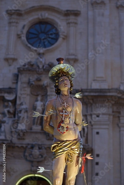 Fototapeta Saint Sebastian Pierced by Arrows on Processional Vara with Illuminated Baroque Church in the Background.
Solemn Night Procession with the Statue of Saint Sebastian of Ferla.