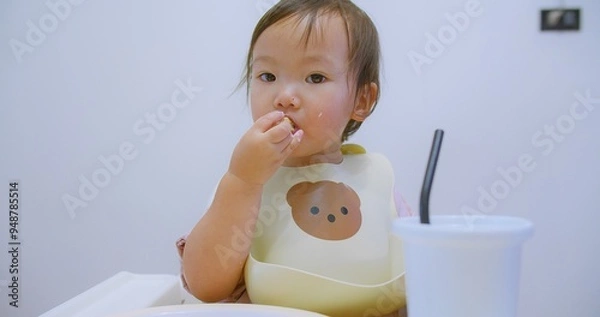 Obraz Cute toddler enjoying eating a snack in a high chair wearing a bear-themed bib in a bright and modern kitchen, Perfect for family, childhood, and home lifestyle BLW themes.