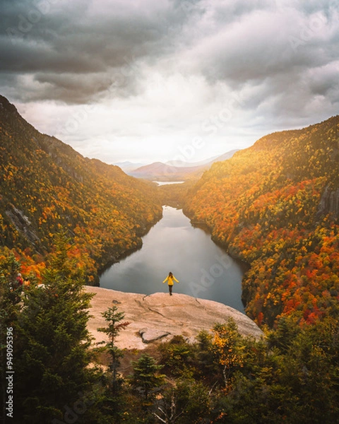 Fototapeta Epic Autumn Indian Head Adirondacks Viewpoint Female Hiker Yellow Jacket Stands in Sun Rays on Mountain. Colorful Fall Foliage and Lake Powerful Scene