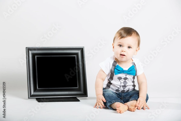 Fototapeta a cute 1 year old baby sits next to a blank black picture frame in a white studio setting. The boy is looking at the camera curiously. He is dressed in Tshirt, jeans, suspenders and blue bow tie